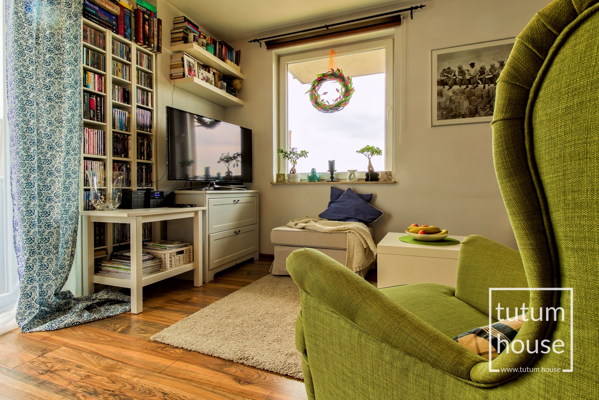 Przytulny salon z zielonym fotelem, regałem pełnym książek i dekoracyjnym wieńcem w oknie, podkreślający ciepłą atmosferę wnętrza.