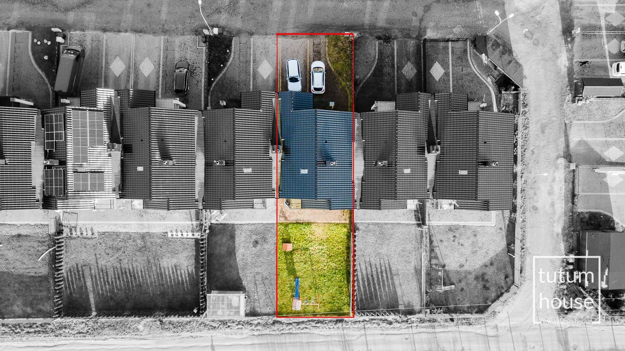 Zdjęcie z lotu ptaka przedstawiające działkę z domem w zabudowie szeregowej, z dwoma zaparkowanymi samochodami na podjeździe i ogrodem z huśtawką, otoczoną przez inne domy, w większości w czerni...