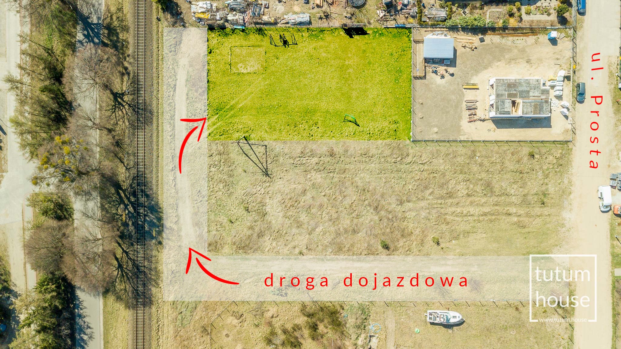 Zdjęcie z drona przedstawiające działkę budowlaną z zaznaczoną drogą dojazdową, w tle widoczny szkielet domu w trakcie budowy, obok torów kolejowych.