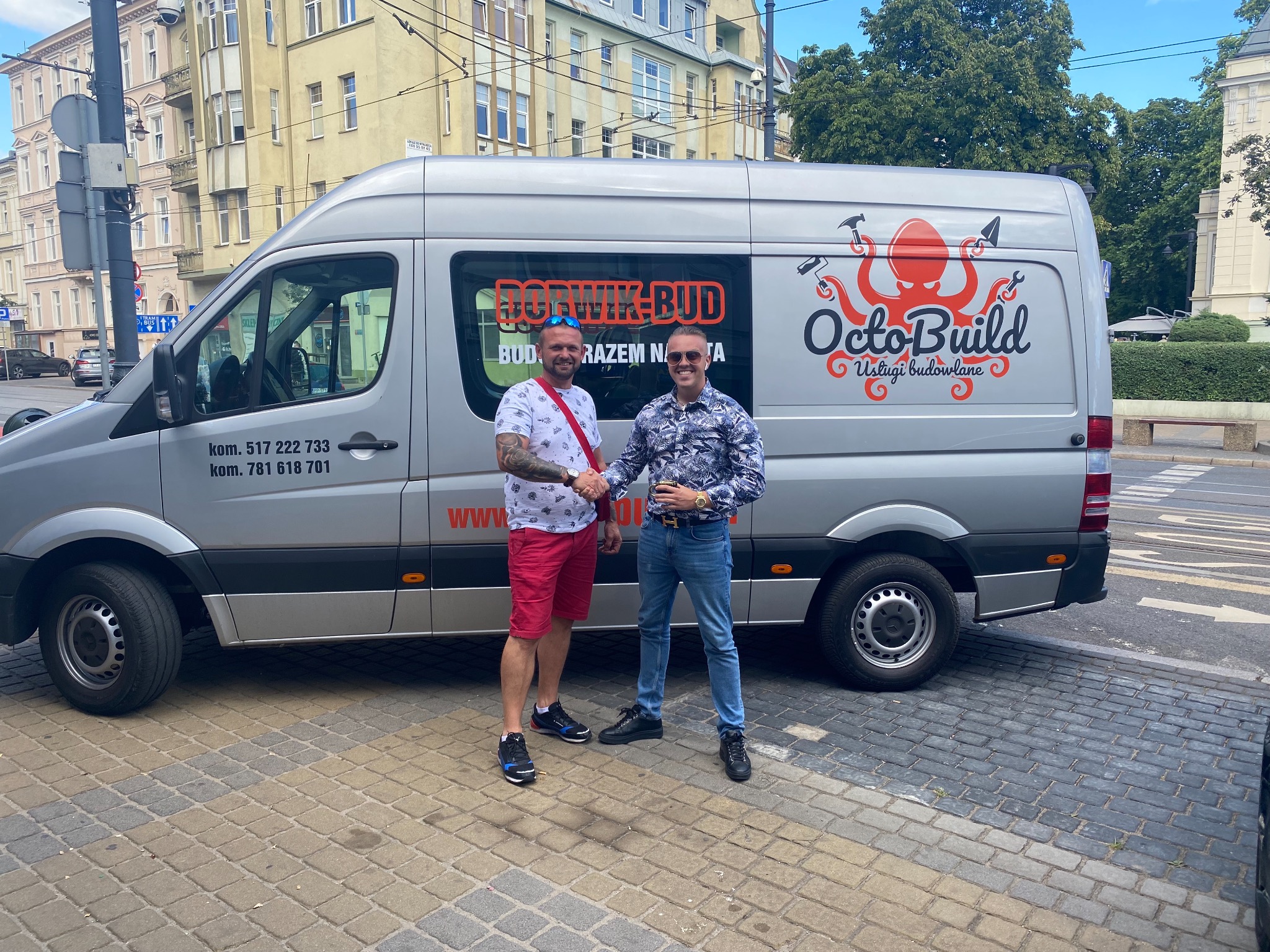 Dwóch mężczyzn w okularach ściska sobie dłonie na tle srebrnego busa z logo firmy budowlanej OctoBuild, zaparkowanego przy ulicy w słoneczny dzień.