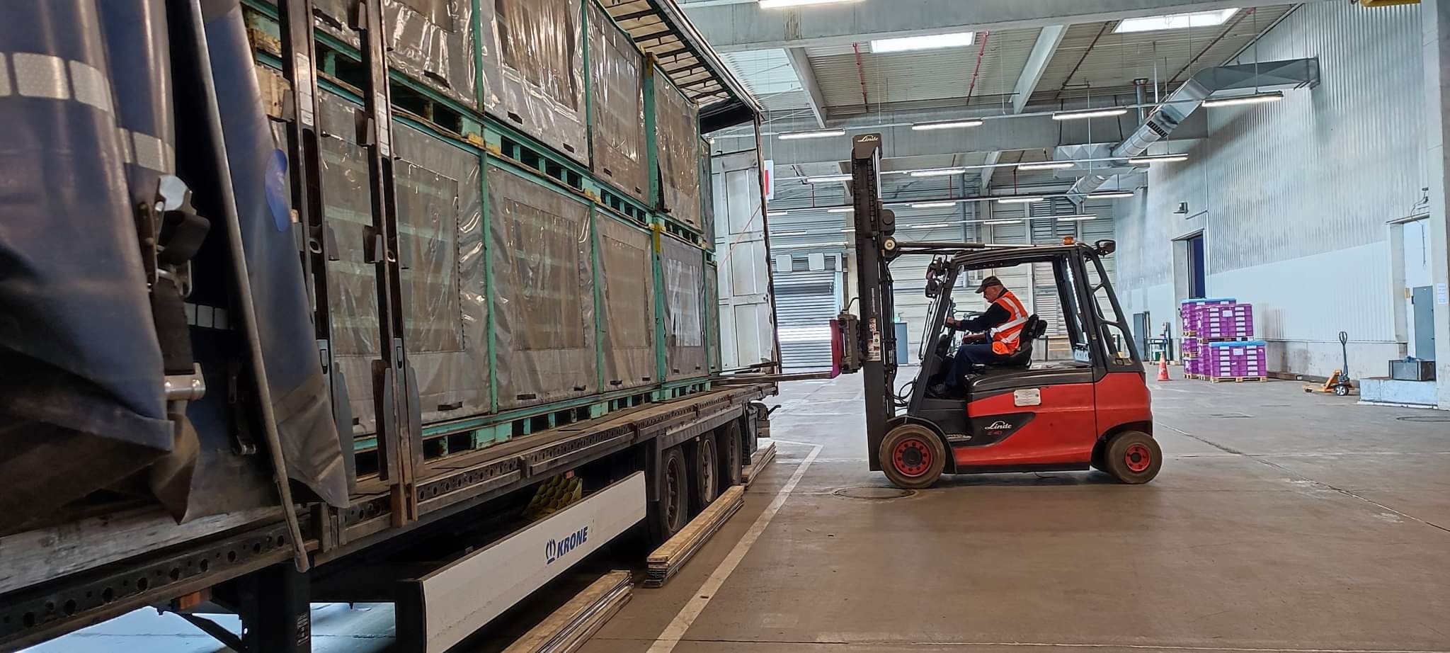 Wózek widłowy Linde z operatorem w kamizelce odblaskowej ładuje towar w metalowych kontenerach na naczepę ciężarówki w magazynie.