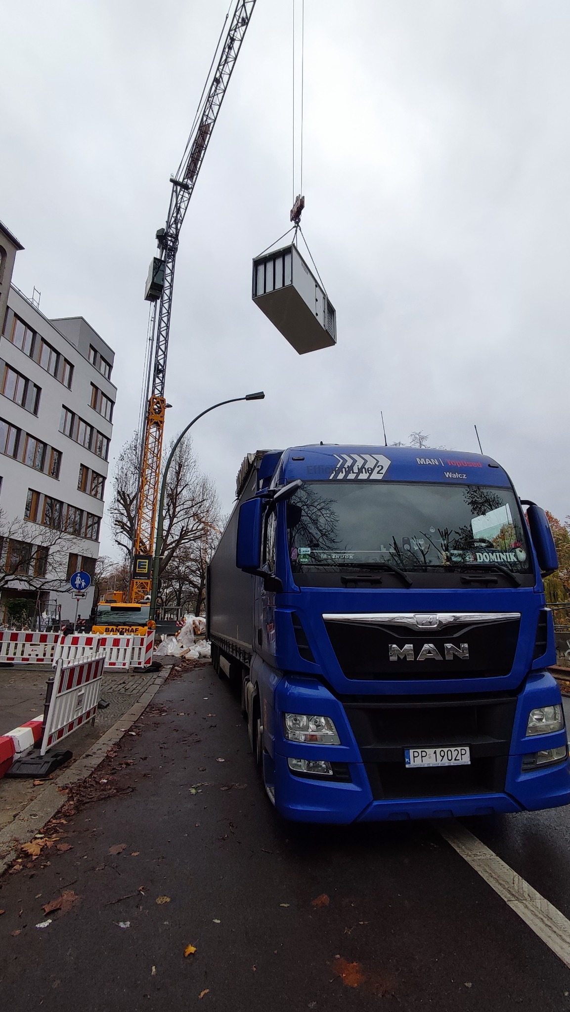 Niebieski ciągnik siodłowy MAN z naczepą na tle miejskiej ulicy podczas podnoszenia kontenera przez pomarańczowy dźwig budowlany, widoczne zabezpieczenia placu budowy.