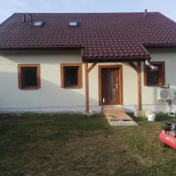 Dom jednorodzinny z brązowym dachem z dachówki, drewnianą ramą okienną i nowym wejściem z podestem. Na trawniku widoczny kompresor i wiadro z farbą.