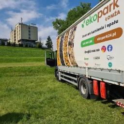 Ciężarówka z reklamą firmy EkoPark Łomża na tle zielonego wzgórza i budynku. Reklama przedstawia drewno opałowe i kamień.