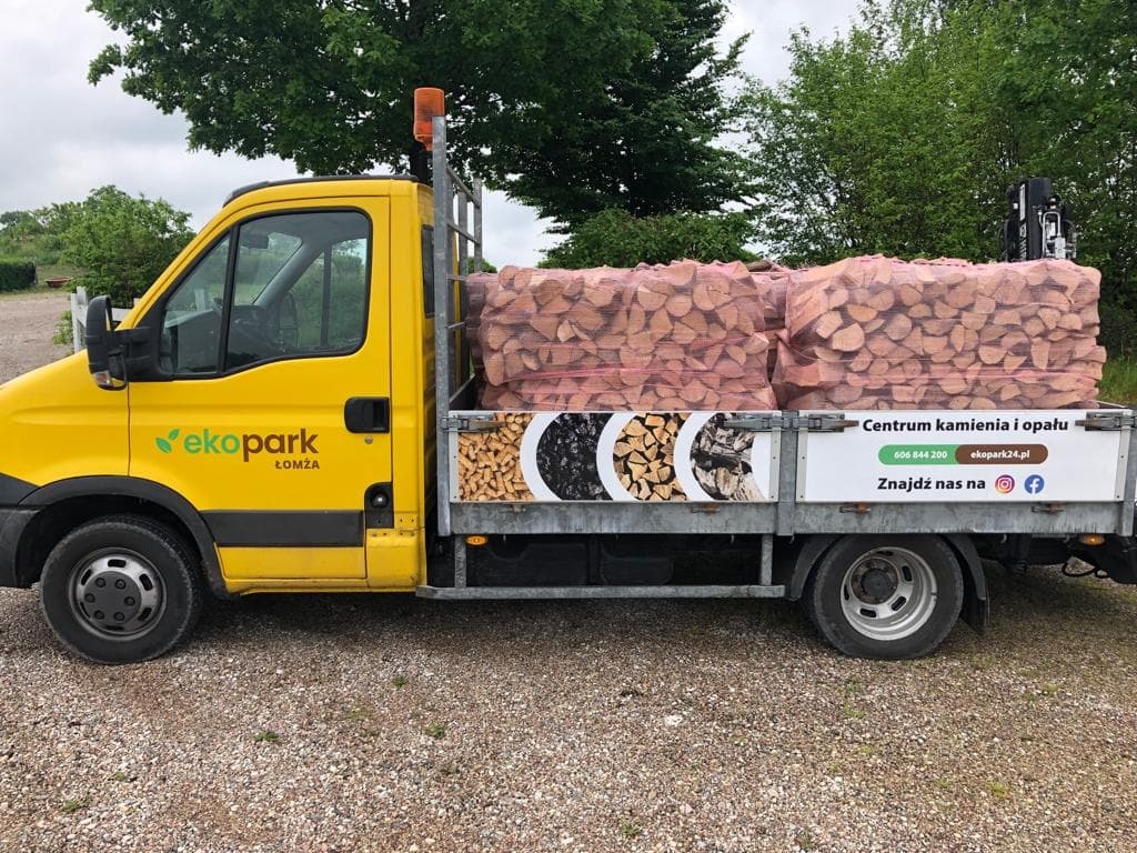 Żółty samochód dostawczy firmy EkoPark Łomża z ładunkiem drewna kominkowego w siatkach, zaparkowany na żwirowym podłożu. Na burcie reklama firmy 'Centrum kamienia i opału' z numerem telefonu...