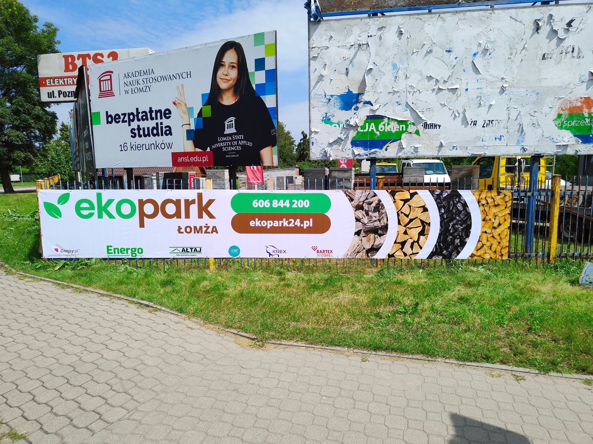 Billboard reklamowy firmy Ekopark Łomża prezentujący drewno opałowe, w tle inne billboardy i drzewa.