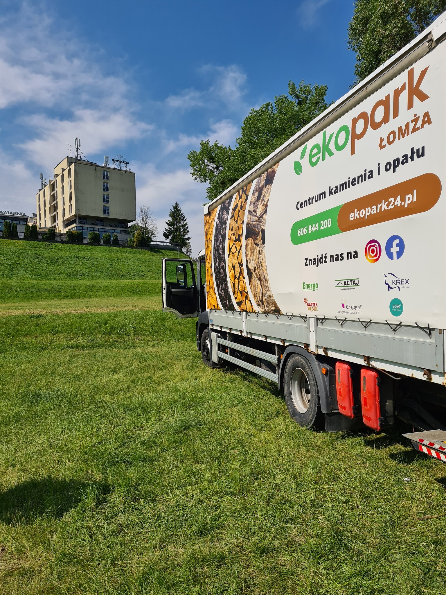 Ciężarówka z reklamą firmy EkoPark Łomża na tle zielonego wzgórza i budynku. Reklama przedstawia drewno opałowe i kamień.
