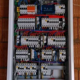 Rozdzielnica elektryczna w obudowie, zamontowana na ścianie z cegły, z widocznymi przewodami i zabezpieczeniami, częściowo zakryta tynkiem w górnej części.