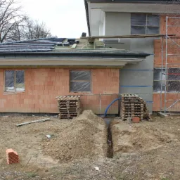 Budowa domu jednorodzinnego w trakcie realizacji: widoczny wykop w ziemi, palety z cegłami, fragment dachu z nową dachówką i rusztowanie przy elewacji.