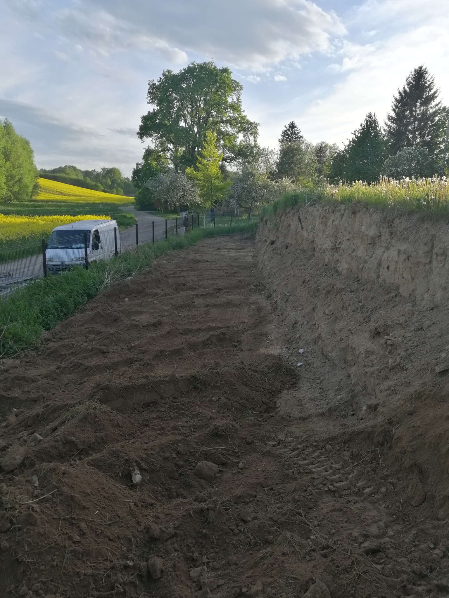 Świeżo wyrównany teren z widocznymi śladami opon, obok ogrodzenie z metalowej siatki, za nim zaparkowany biały bus i pole rzepaku w oddali, po prawej stronie skarpa ziemna porośnięta trawą.