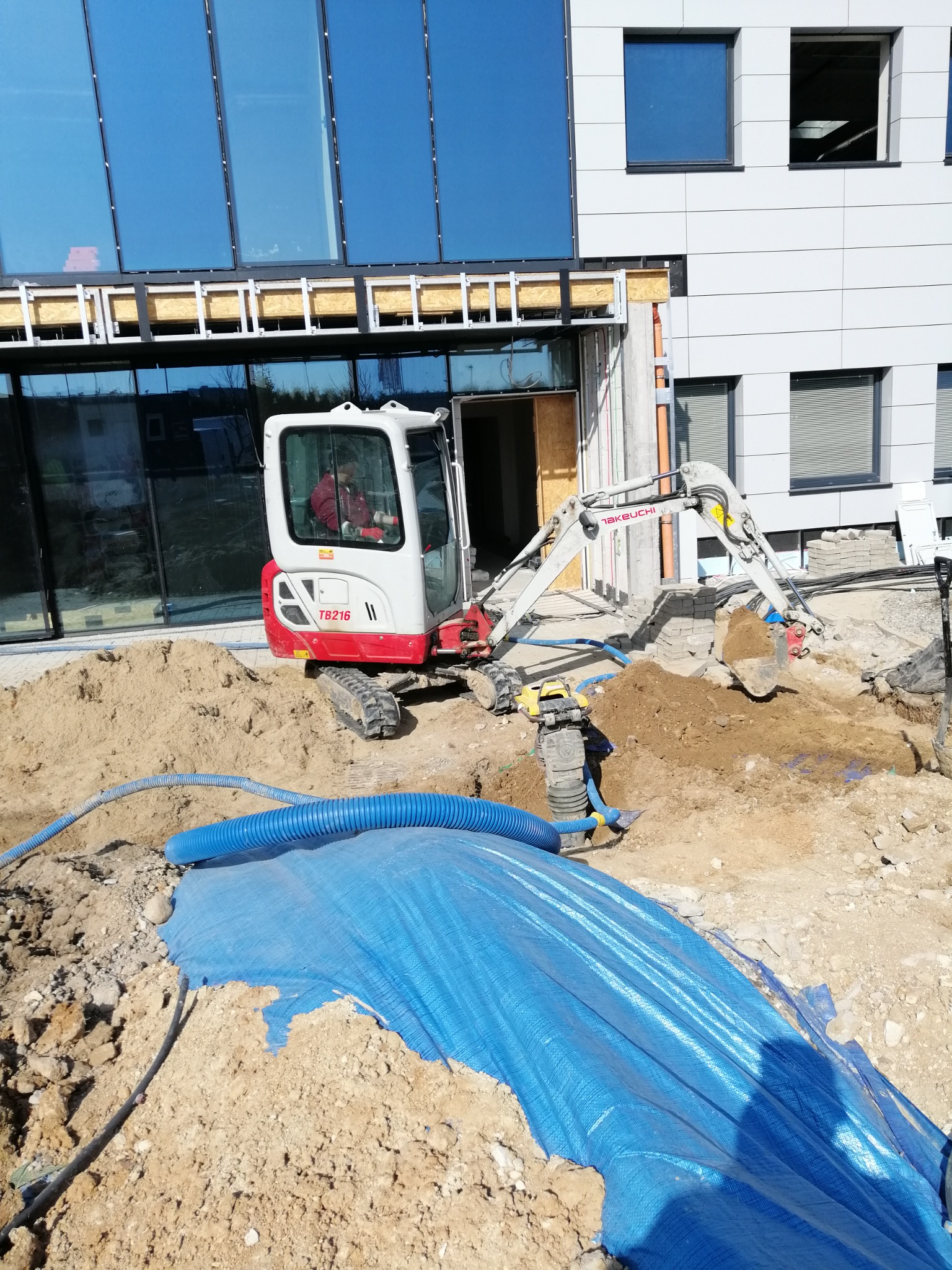 Mini koparka Takeuchi TB216 w trakcie prac ziemnych na placu budowy, obok niebieska plandeka i zagęszczarka wibracyjna, budynek w tle z dużymi oknami.