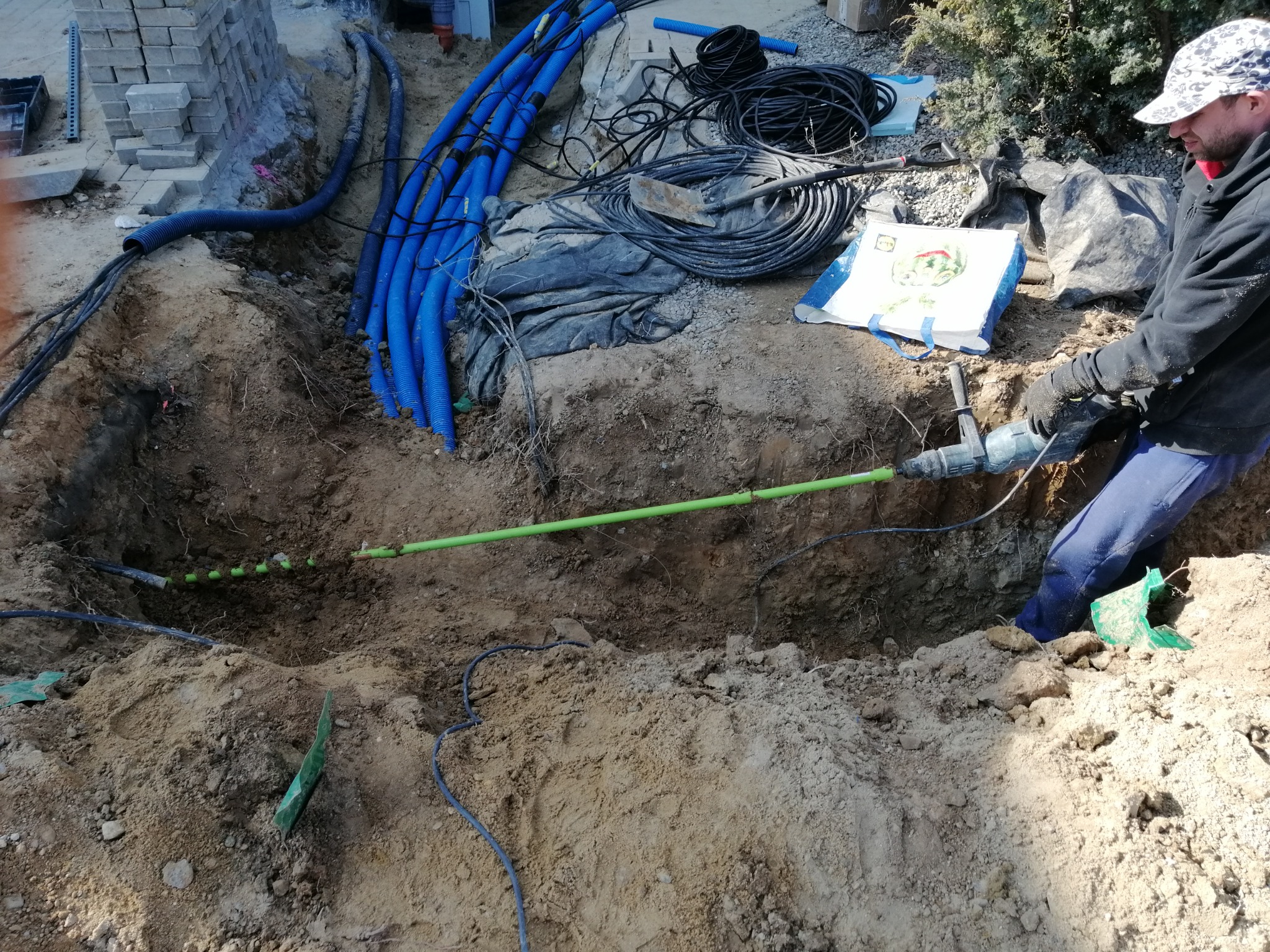 Pracownik w czapce i rękawicach roboczych kopie wykop pod instalacje elektryczne, widoczne niebieskie rury karbowane, kable i zielona rura w ziemi.
