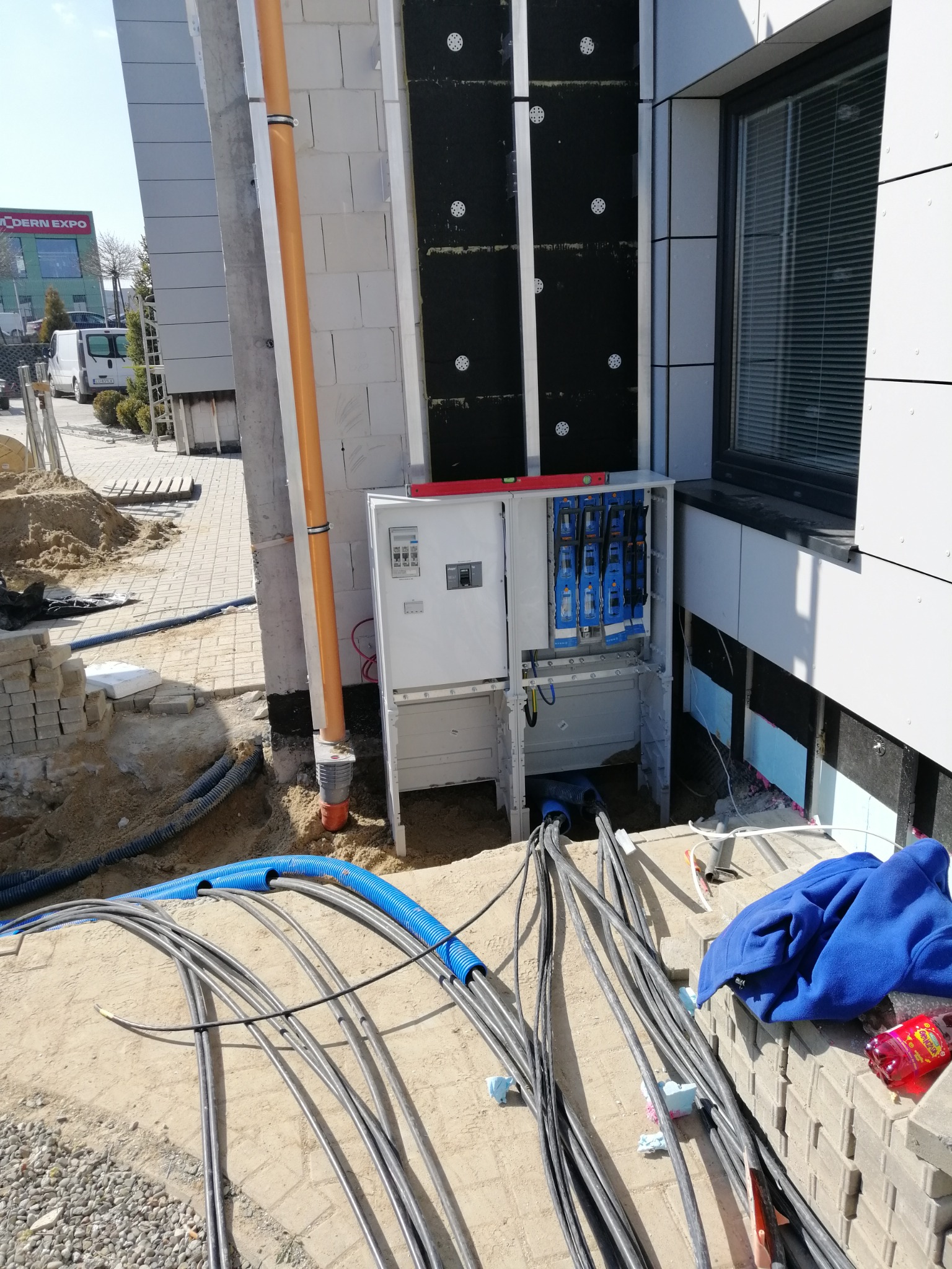 Otwarta skrzynka elektryczna z zabezpieczeniami, poziomica na górze, liczne kable w peszlach i bez, ułożone na ziemi przy budynku w trakcie budowy.