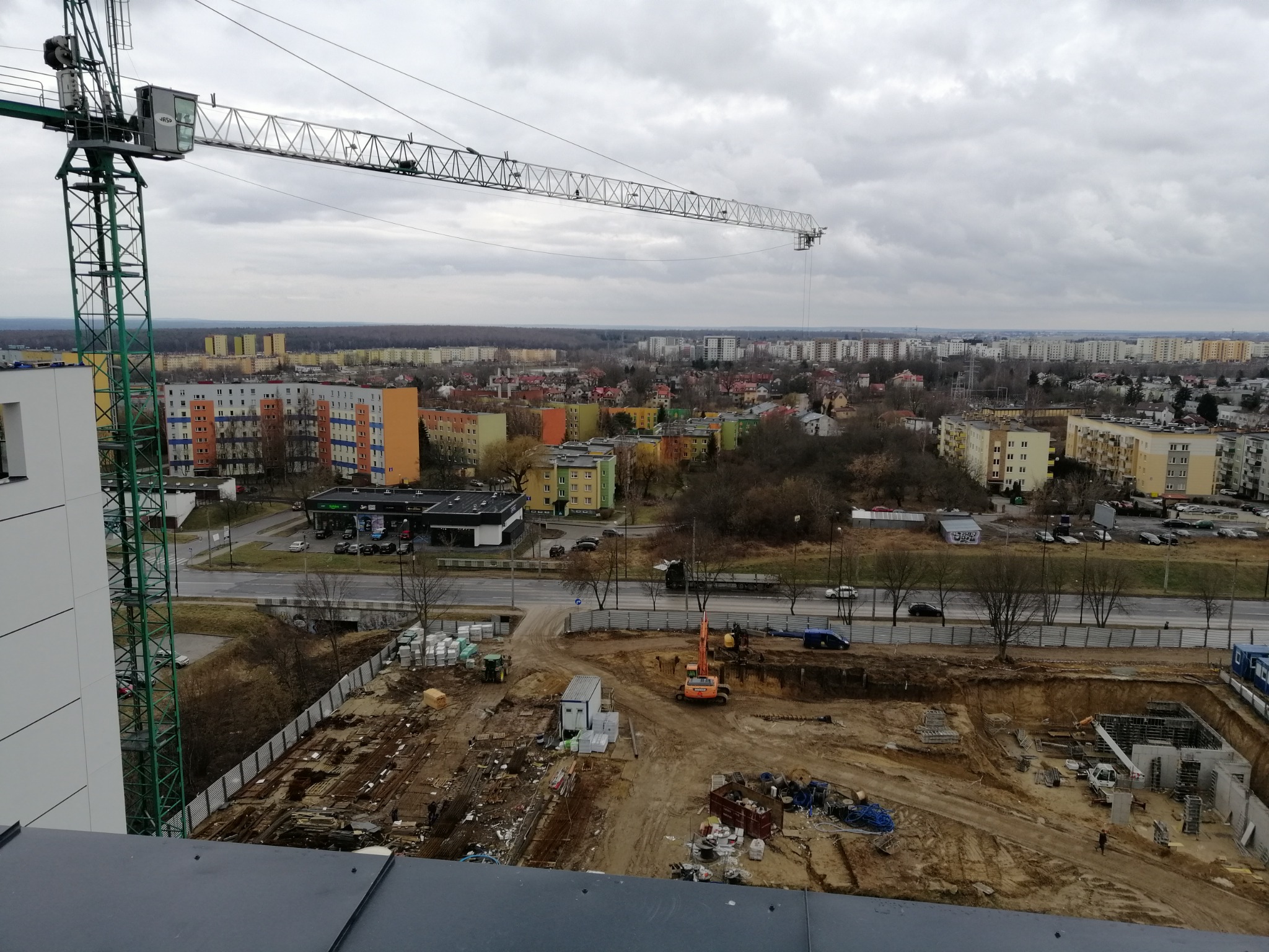 Widok z góry na plac budowy z koparką, żurawiem wieżowym i wykopem pod fundamenty, na tle zabudowy miejskiej w pochmurny dzień.