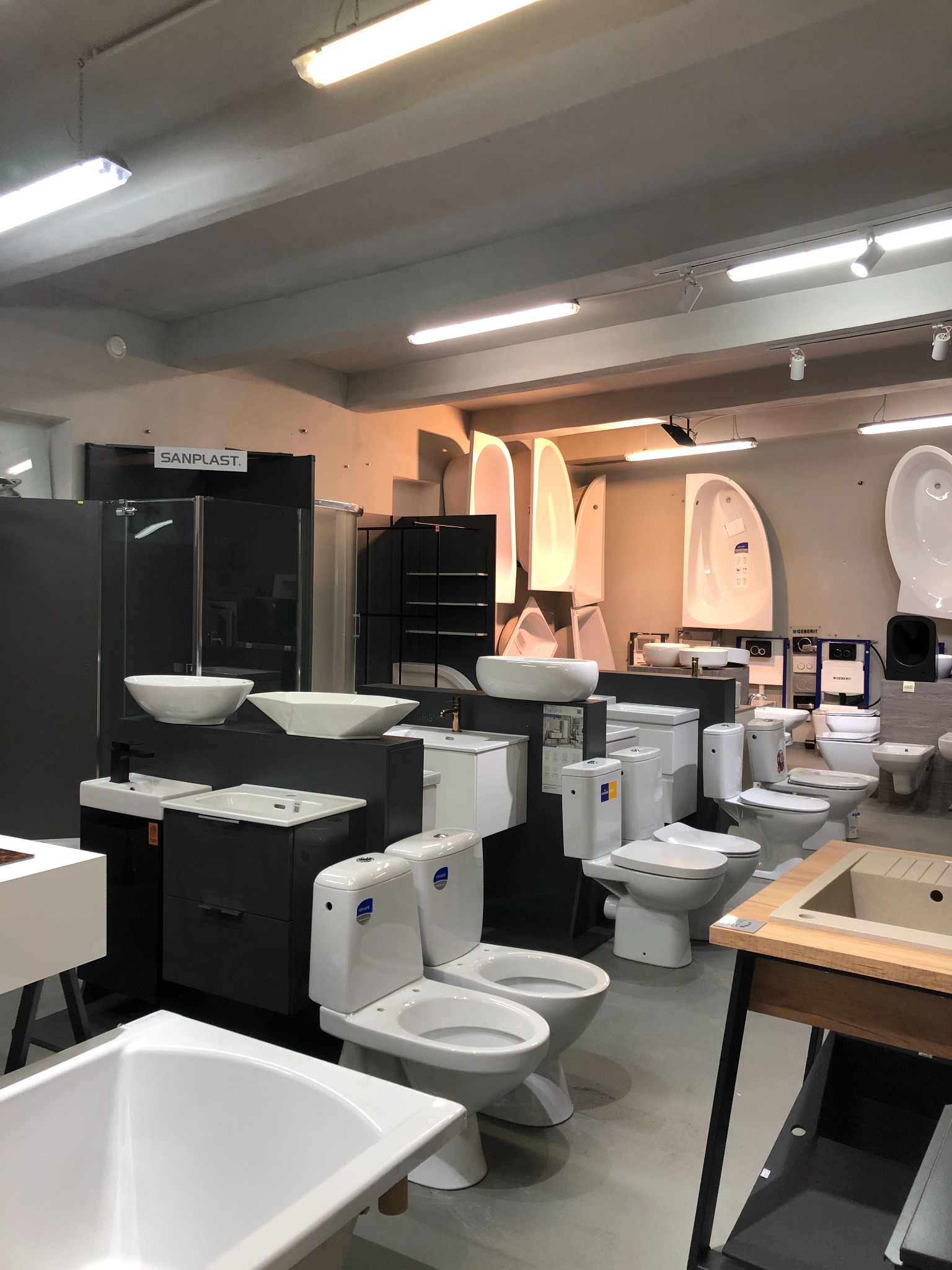 Wystawa w salonie z armaturą łazienkową, prezentująca wanny, umywalki nablatowe o geometrycznych kształtach, kabiny prysznicowe z czarnymi profilami i toalety kompaktowe, oświetlona białymi lampami...