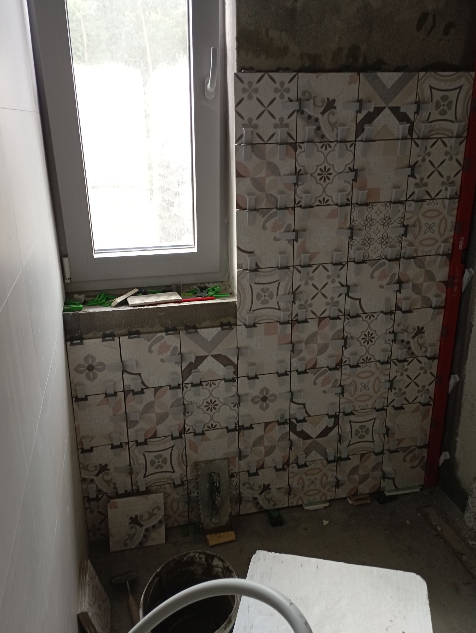 Układanie dekoracyjnych płytek ceramicznych na ścianie w pobliżu okna, widoczne narzędzia glazurnicze i poziomica, etap prac wykończeniowych.