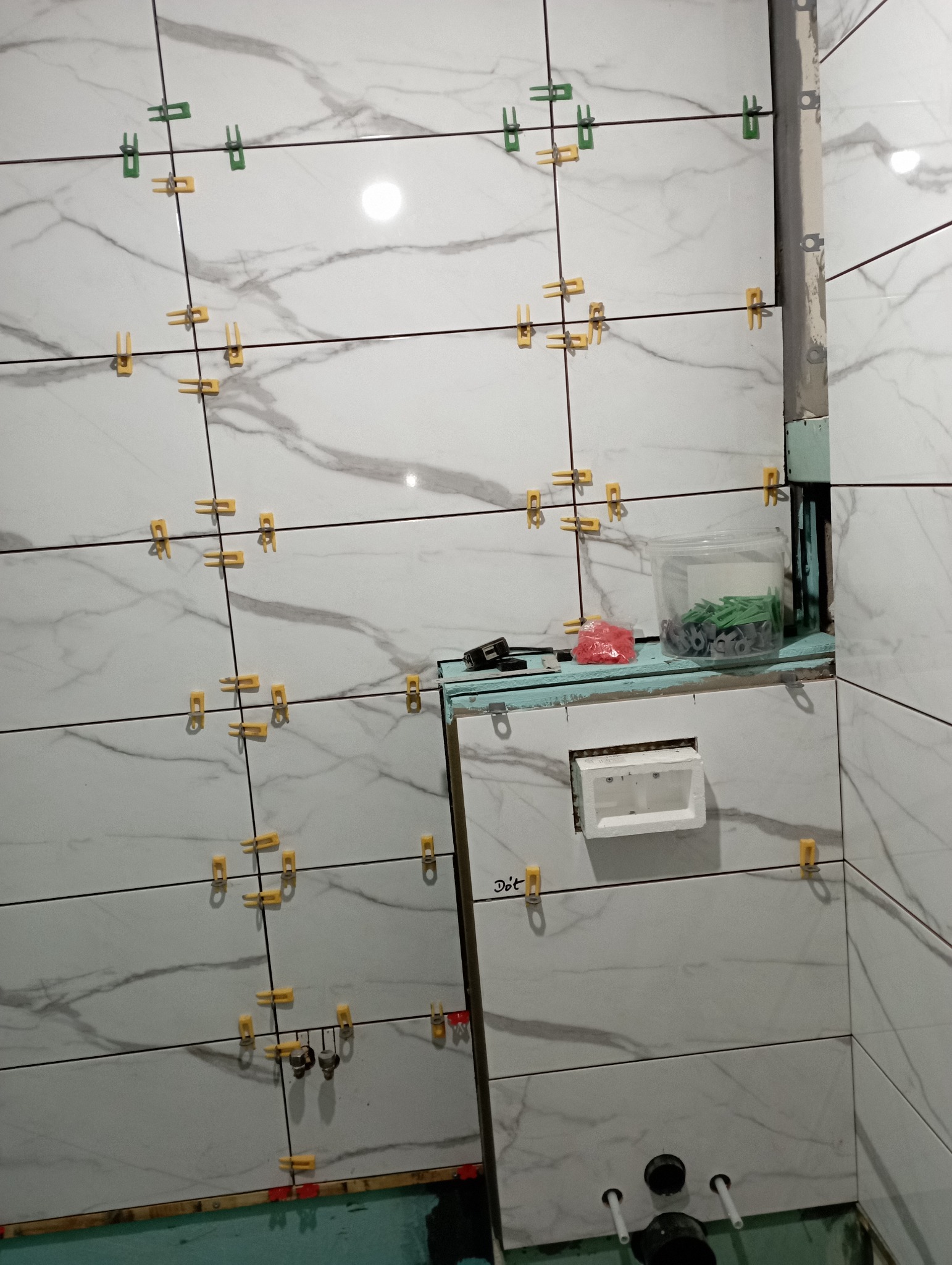 Układanie białych płytek imitujących marmur z użyciem żółtych i zielonych klipsów poziomujących, widoczna instalacja podtynkowa WC, etap prac wykończeniowych w łazience.