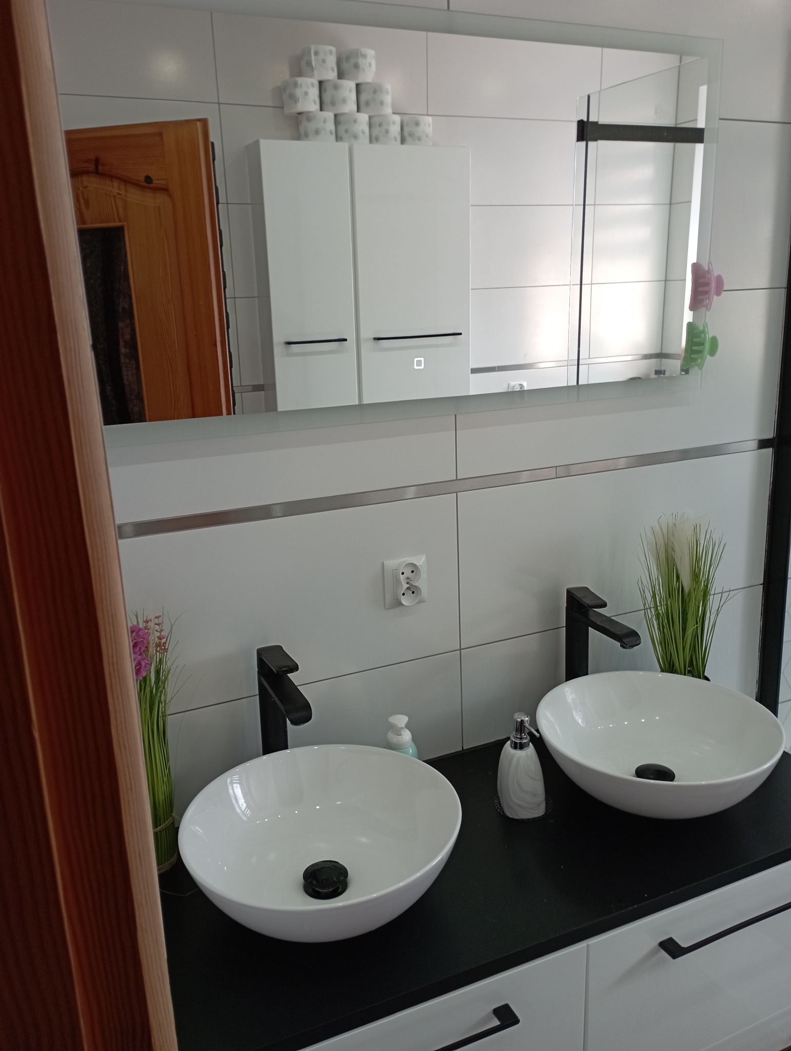 Nowoczesna łazienka z dwoma okrągłymi umywalkami nablatowymi, czarnymi bateriami, białymi szafkami z minimalistycznymi uchwytami i lustrem, na którym odbija się fragment drewnianych drzwi...