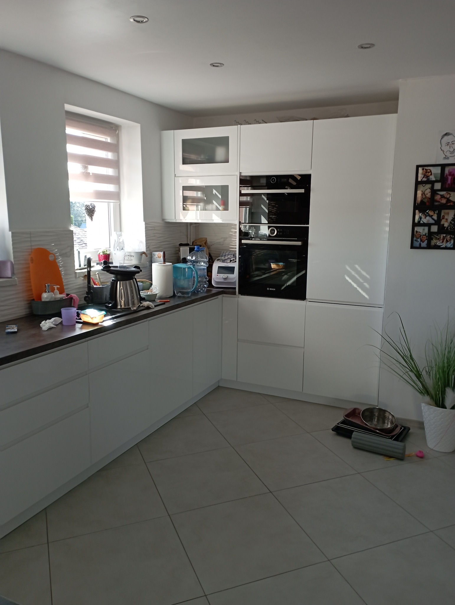 Nowoczesna kuchnia z białymi szafkami, ciemnym blatem i beżową podłogą. Na blacie widoczne akcesoria kuchenne, w tym czajnik, filtry do wody i pojemniki. Na ścianie wisi kolaż zdjęć rodzinnych.