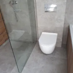 Nowoczesna łazienka z szarą, teksturowaną glazurą, kabiną prysznicową typu walk-in z odpływem liniowym i podwieszaną miską WC z chromowanym przyciskiem spłukującym.