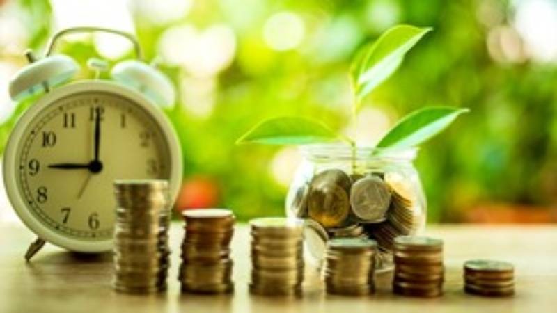 Stos monet ułożony w rosnące kolumny obok białego budzika z godziną 9:00, a w tle słoik wypełniony monetami z młodą rośliną, symbolizujący wzrost finansowy i planowanie czasu.