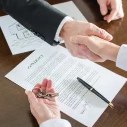 Uścisk dłoni nad podpisaną umową, klucze w dłoni, schemat domu w tle, eleganckie pióro obok kontraktu.