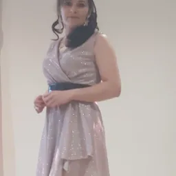 Kobieta w błyszczącej, asymetrycznej sukience z czarnym paskiem, pozująca na jasnym tle.