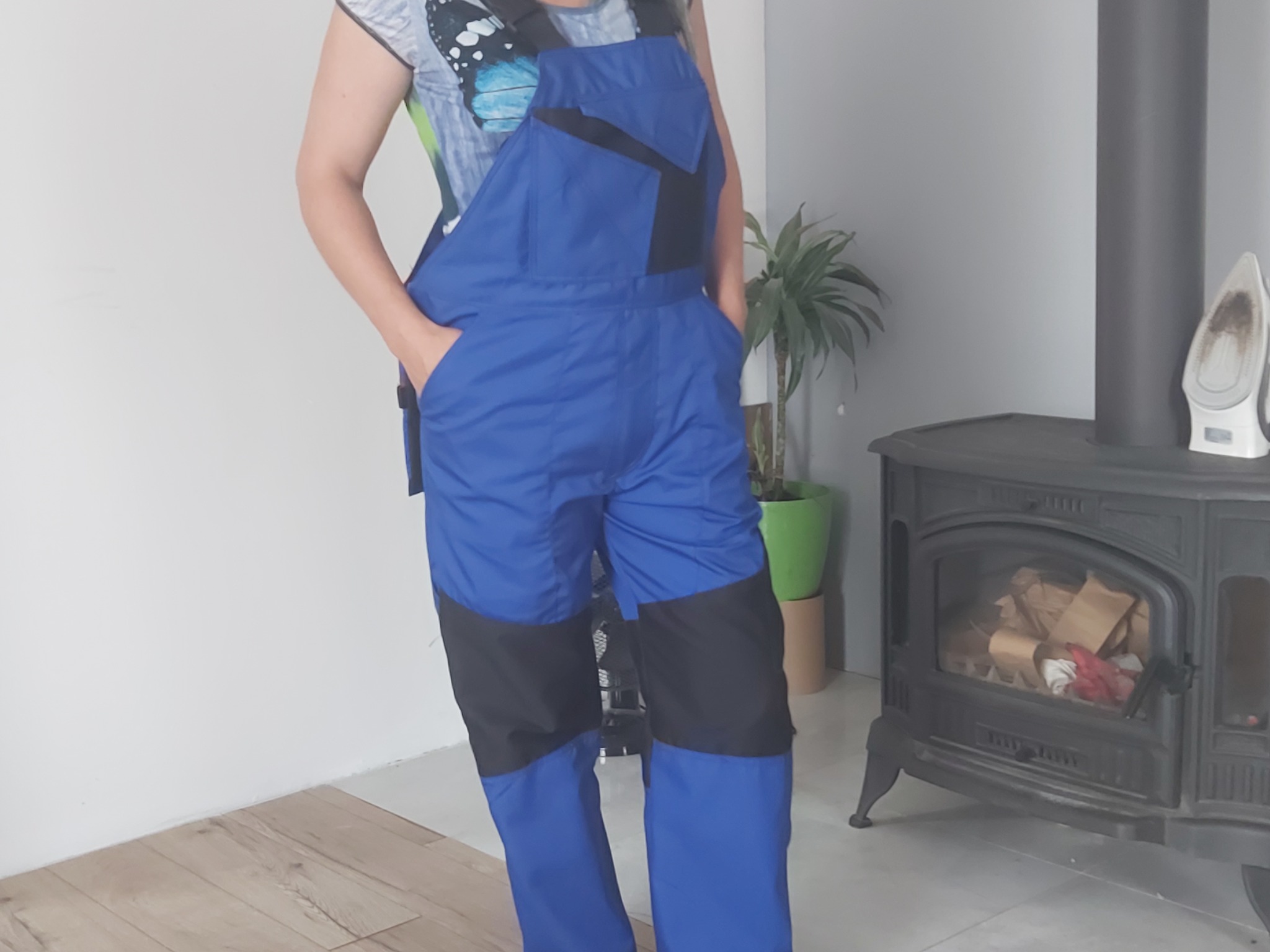 Osoba w niebieskich roboczych ogrodniczkach z czarnymi wstawkami, stojąca w pomieszczeniu z piecem kaflowym i rośliną doniczkową w tle.