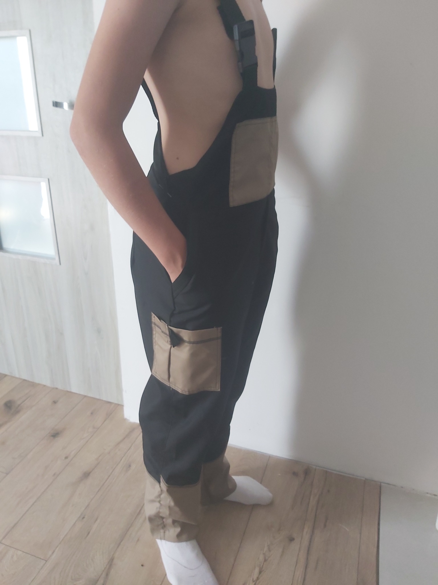 Osoba pozuje w czarnych ogrodniczkach z beżowymi kieszeniami i nogawkami, widoczne białe skarpetki i drewniana podłoga.