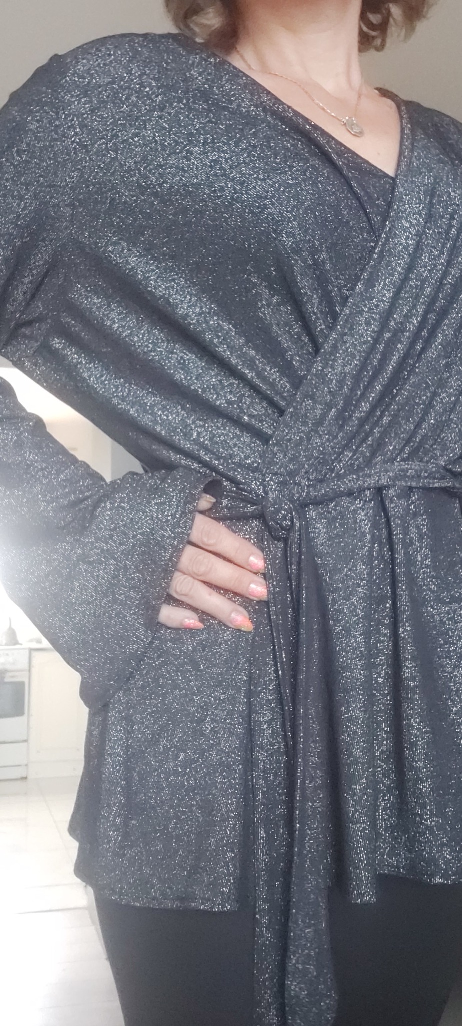 Błyszcząca, grafitowa bluzka z wiązaniem w talii, prezentowana na osobie, z widoczną dłonią z kolorowym manicure.