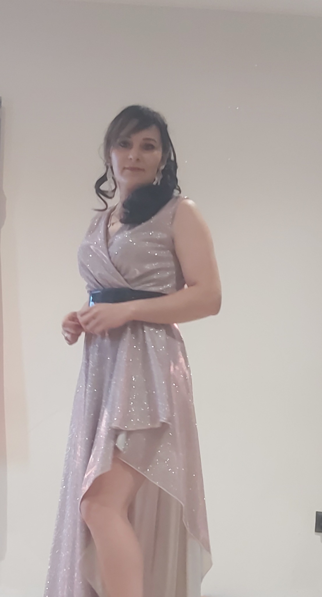 Kobieta w błyszczącej, asymetrycznej sukience z czarnym paskiem, pozująca na jasnym tle.