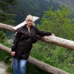 Kobieta w brązowej kurtce i jeansach opiera się o drewnianą barierkę na tle górskiego krajobrazu z jeziorem i lasem.