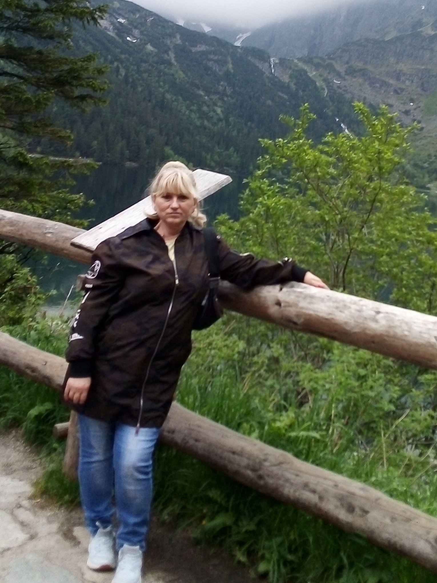 Kobieta w brązowej kurtce i jeansach opiera się o drewnianą barierkę na tle górskiego krajobrazu z jeziorem i lasem.