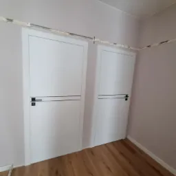 Dwa białe, bezprzylgowe skrzydła drzwiowe z czarnymi poziomymi akcentami i klamkami, zamontowane w ścianie, widoczne ślady pianki montażowej na ościeżnicach i przy suficie.