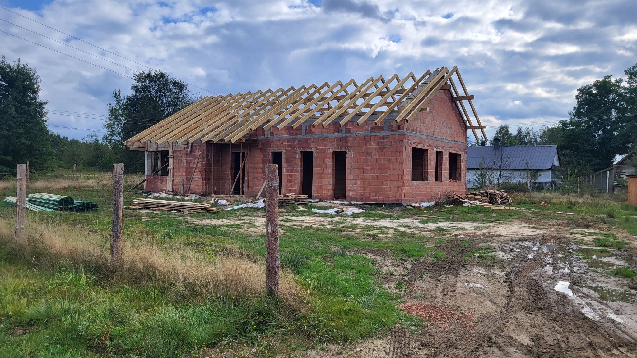 Surowy, ceglany dom w trakcie budowy z drewnianą konstrukcją dachu, otoczony błotnistym terenem i częściowo zarośnięty trawą, pod pochmurnym niebem.