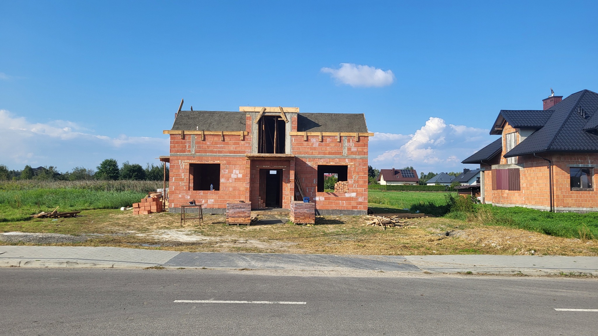 Surowy stan dwukondygnacyjnego domu z czerwonej cegły, z widocznymi otworami okiennymi i drzwiowymi, na tle błękitnego nieba z pojedynczymi obłokami.