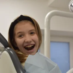 Uśmiechnięta nastolatka z aparatem ortodontycznym w fotelu dentystycznym w Łodzi, widoczna lampa stomatologiczna.