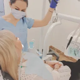 Pacjentka w trakcie zabiegu stomatologicznym; dentystka w niebieskim uniformie reguluje lampę nad głową pacjentki, widoczne narzędzia i akcesoria dentystyczne.