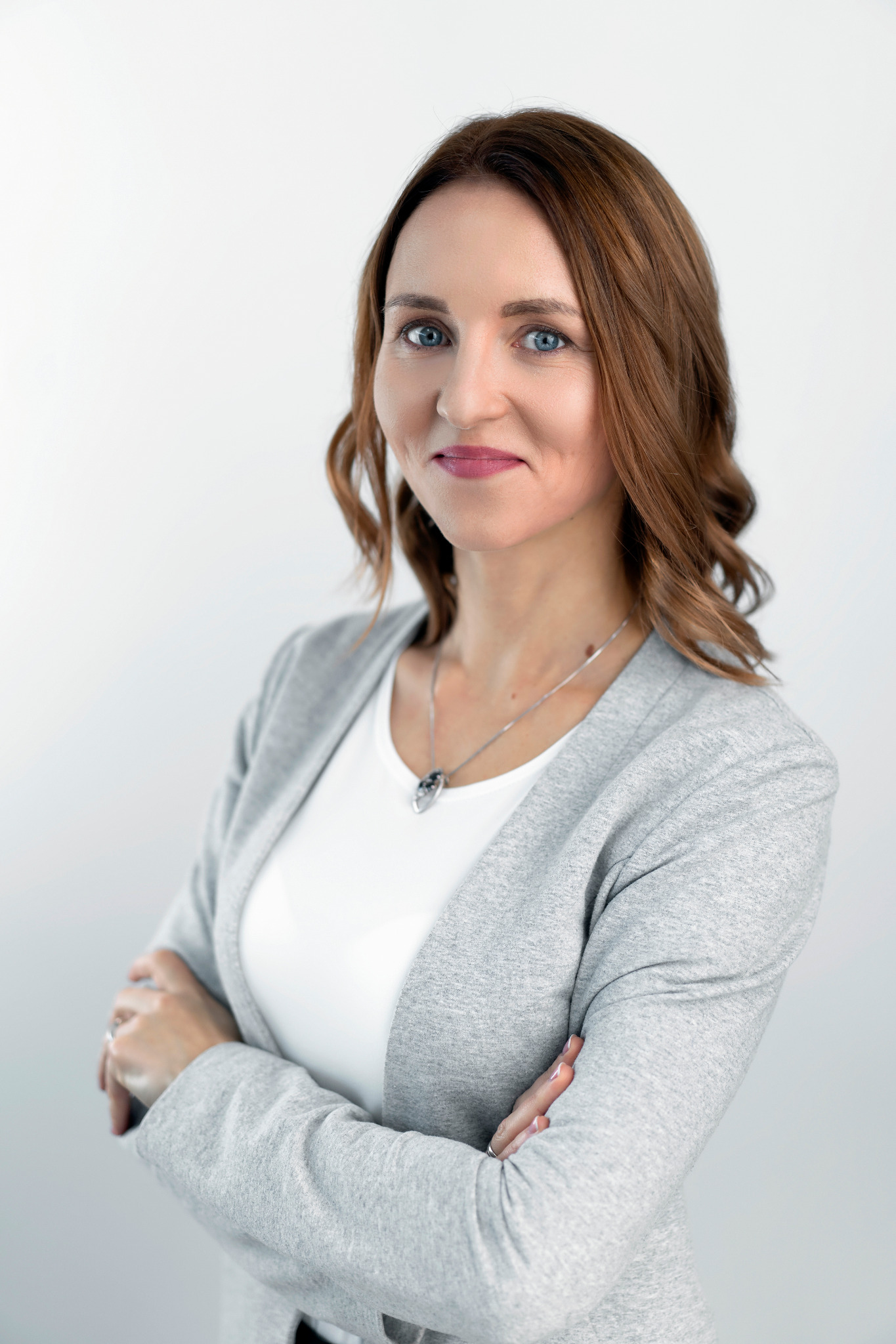 Portret uśmiechniętej kobiety z rudymi włosami i niebieskimi oczami, ubrana w szary kardigan i białą bluzkę, z rękami skrzyżowanymi na piersiach, na jasnym tle.