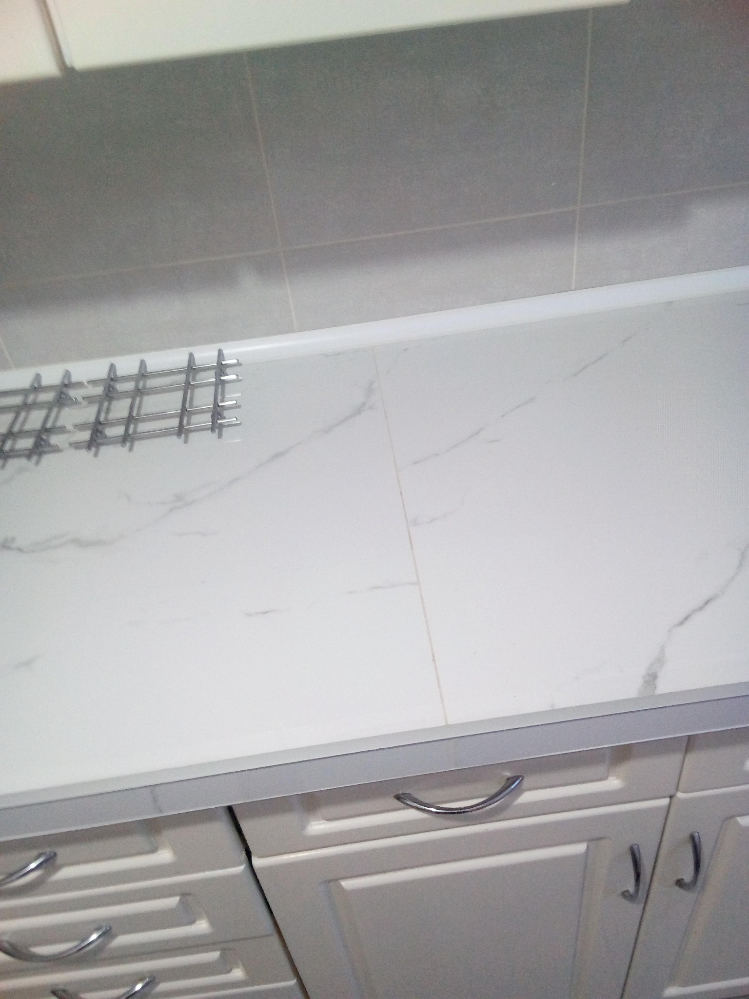 Biały blat z marmurowym wzorem w łazience, widoczna suszarka na naczynia i szafki pod umywalką z chromowanymi uchwytami.