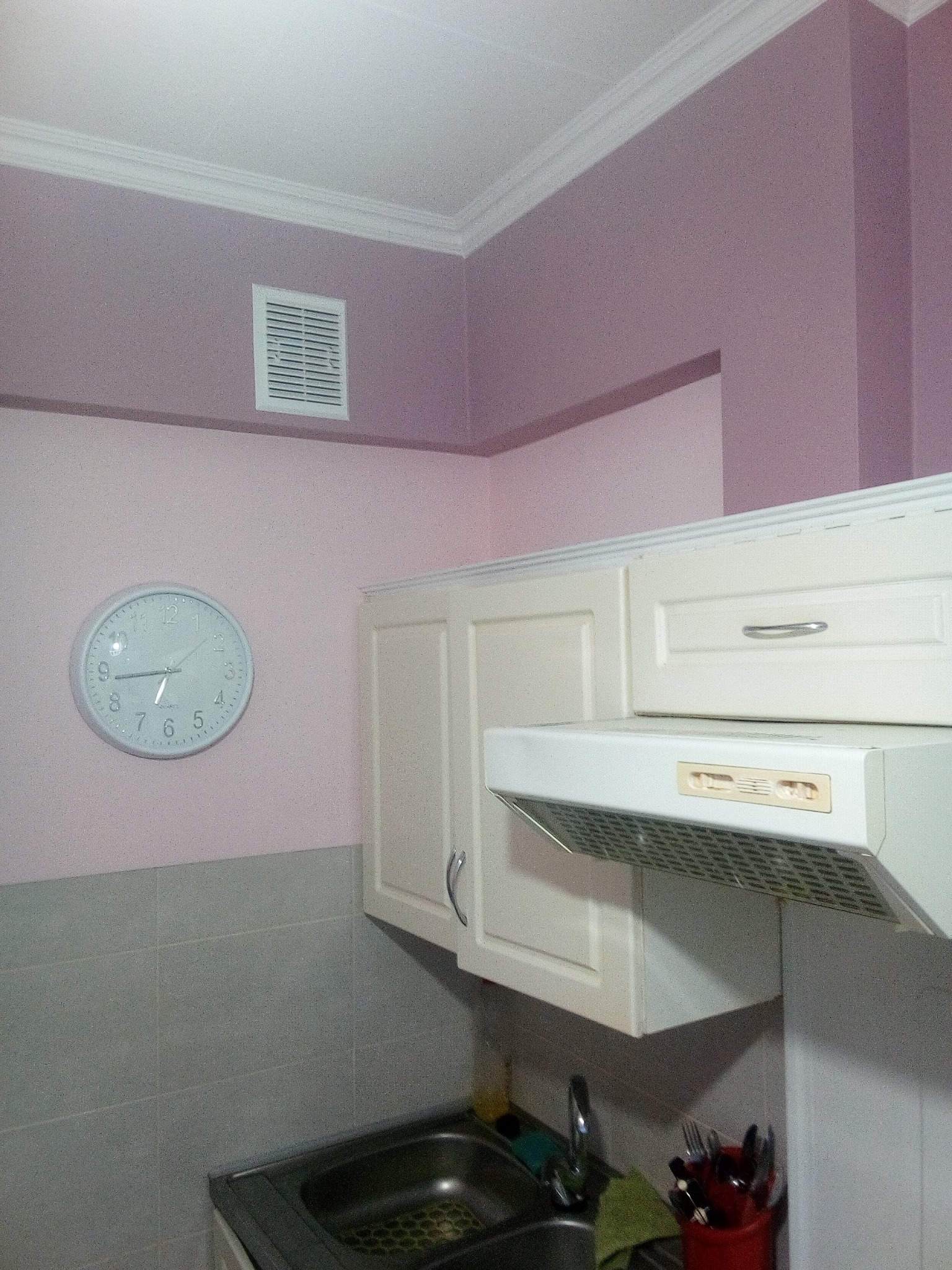Jasna kuchnia z szafkami wiszącymi, okapem i szarymi płytkami nad zlewem, ściany pomalowane na jasny fiolet, na ścianie okrągły zegar.