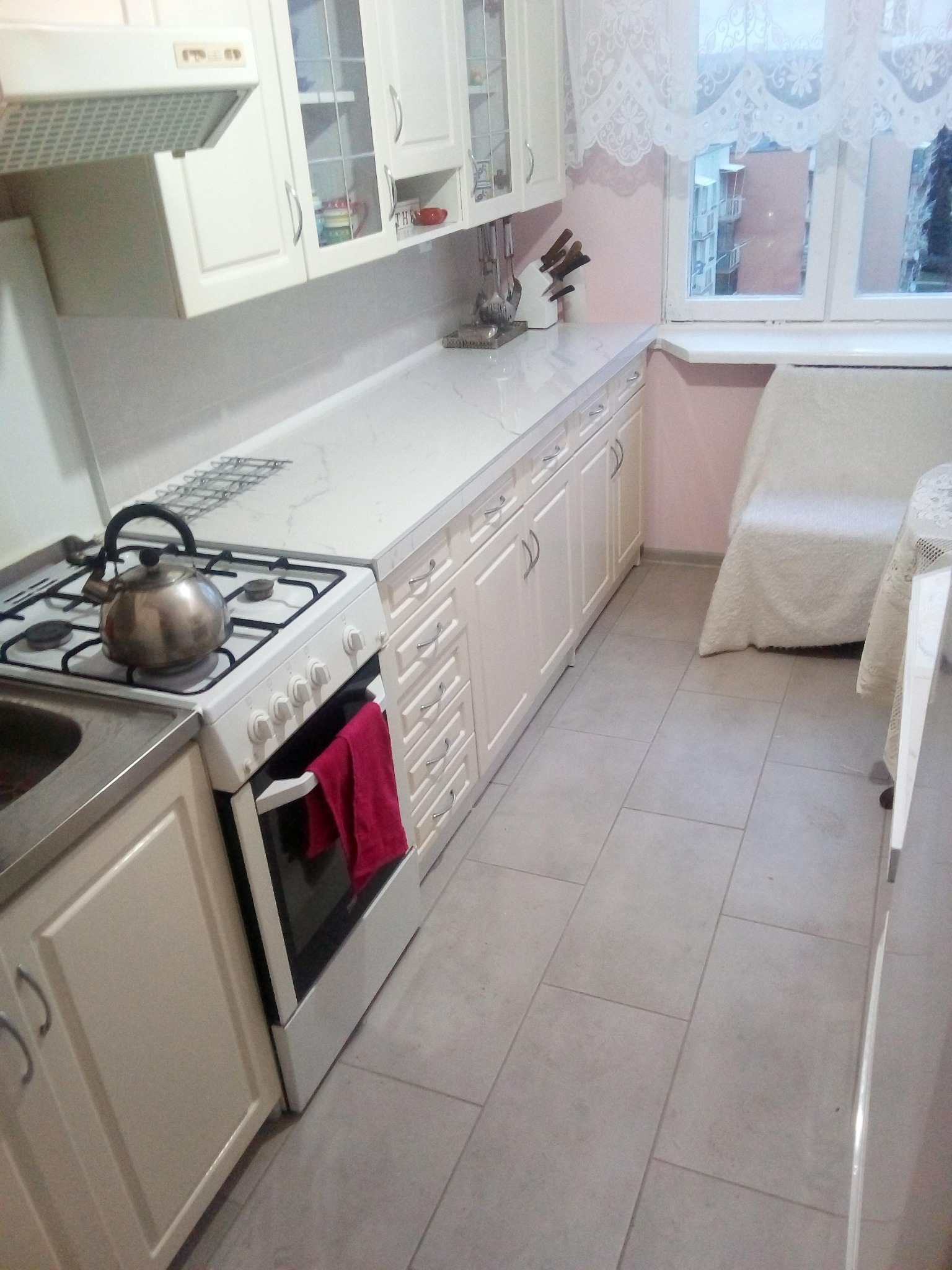 Jasna, funkcjonalna kuchnia z białymi szafkami, marmurowym blatem, czajnikiem na płycie gazowej i czerwonym ręcznikiem na piekarniku.