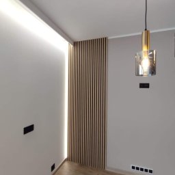 Kriss-Bud Kompleksowe usługi budowlane - Panel z drewnianych lameli na ścianie z podświetleniem LED, minimalistyczna lampa wisząca z elementami złota, gładkie ściany w odcieniach szarości, drewnopodobna podłoga, czarne gniazdka elektryczne.