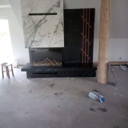 Kriss-Bud Kompleksowe usługi budowlane - Nowoczesny kominek z marmurową obudową i dekoracyjnym panelem z motywem drzewa, surowy pień drzewa jako element wystroju, podłoga w trakcie remontu.