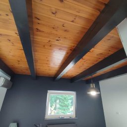Kriss-Bud Kompleksowe usługi budowlane - Drewniany sufit z belkami pomalowanymi na ciemny kolor, szara ściana, okno z widokiem na zieleń i lampa wisząca w stylu industrialnym.