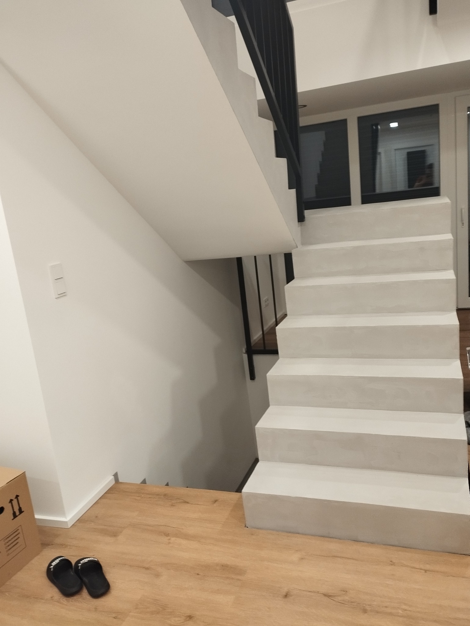 Nowoczesne, białe schody z czarną balustradą w jasnym wnętrzu po remoncie, widoczne drewniane panele podłogowe i kartonowe pudło.