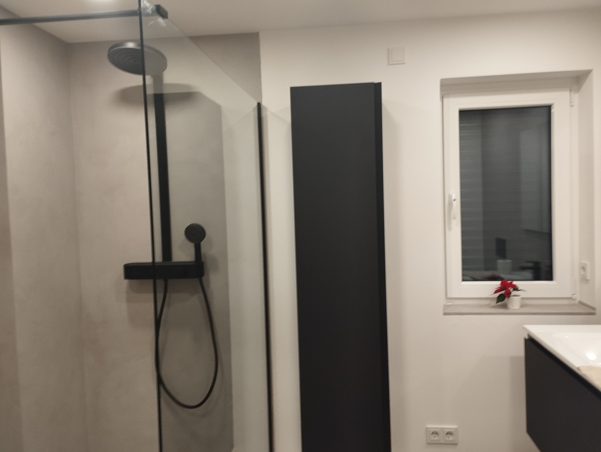 Nowoczesna łazienka z czarną armaturą prysznicową, przeszkloną kabiną, wysoką szafką i oknem z dekoracją kwiatową.