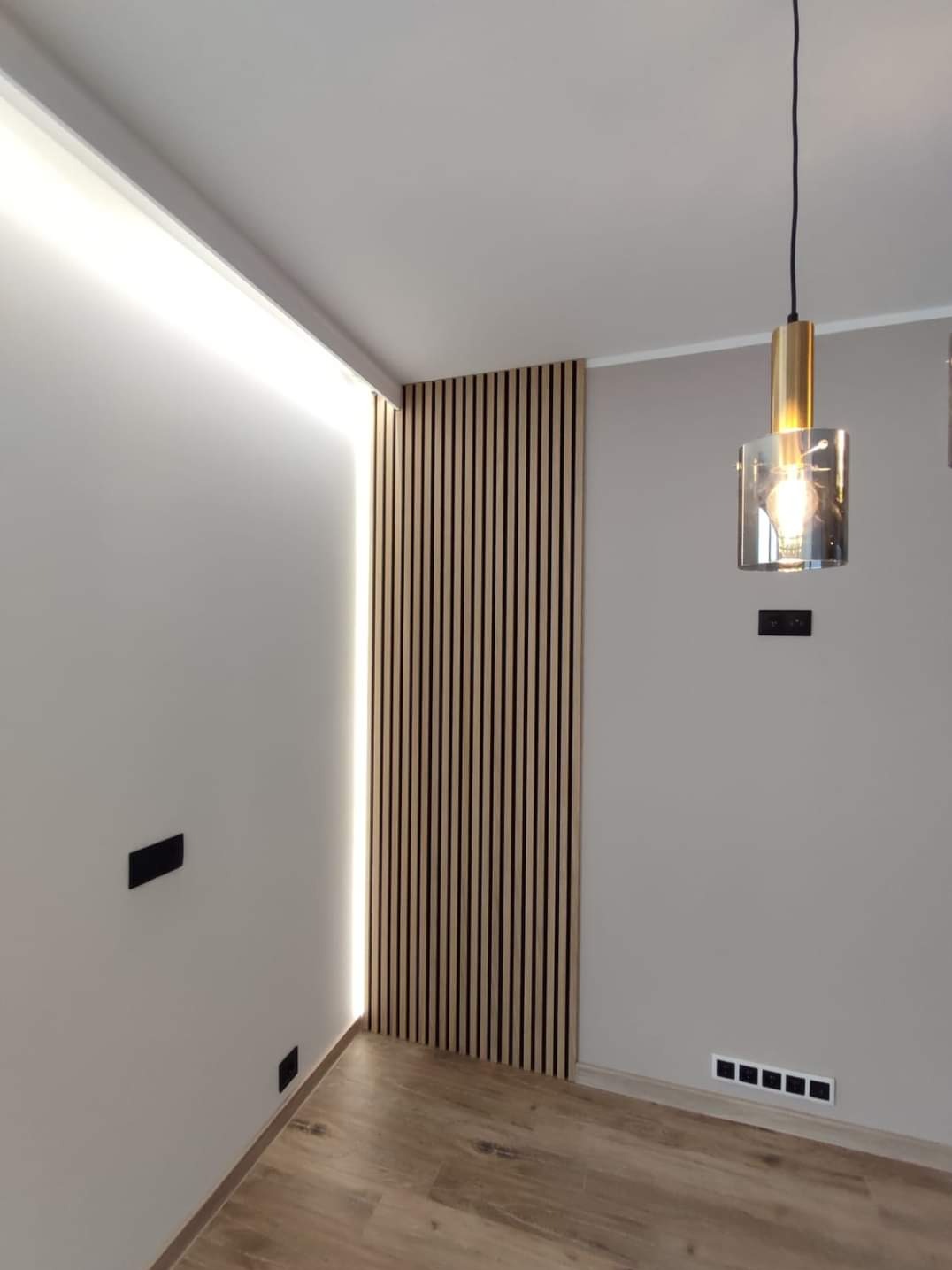 Panel z drewnianych lameli na ścianie z podświetleniem LED, minimalistyczna lampa wisząca z elementami złota, gładkie ściany w odcieniach szarości, drewnopodobna podłoga, czarne gniazdka elektryczne.