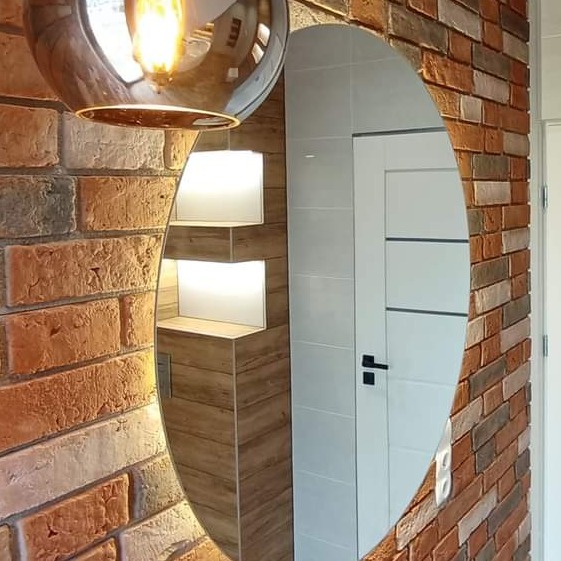 Okrągłe lustro z podświetleniem LED w łazience z ceglaną ścianą i wnęką z półkami, odbijające białe drzwi z czarną klamką.