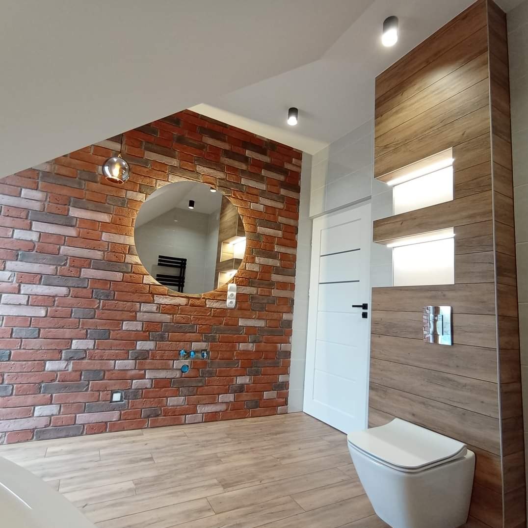 Nowoczesna łazienka z akcentem cegły na ścianie, okrągłe lustro z podświetleniem, drewnopodobne płytki na podłodze i zabudowie toalety.